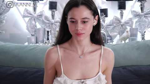 Media: Video of a young, slender woman with fair skin and dark hair, wearing a white lace camisole and small earrings. Background features a bed with silver decorations, including butterfly ornaments.