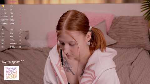 Media: Video of a young woman with fair skin, red hair in a ponytail, and a tattoo on her chest, wearing a white hoodie, sitting on a gray bed with pink pillows.