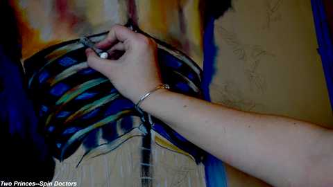 Media: Video of a woman's hand painting a vibrant, abstract, multicolored mural on a beige wall. Her arm, adorned with a silver bracelet, is seen in the foreground.