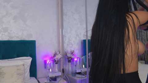 Media: Video of a woman with long, straight black hair, wearing a black top, standing in a modern bedroom with a teal headboard, mirrored walls, and purple LED lights.