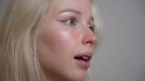 Video of a young, fair-skinned woman with long blonde hair, wearing minimal makeup, featuring prominent eyeliner. She has a slight open-mouth expression, with visible teeth. Background is a plain, light-colored wall.