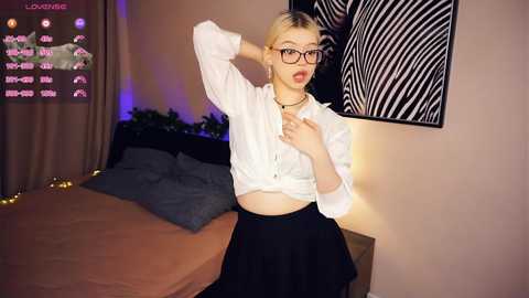 Media: Video of a young, fair-skinned woman with short blonde hair, wearing glasses and a white button-up shirt tied at the waist, black skirt, and a black choker. She poses in a dimly lit bedroom with a bed and a zebra-patterned wall.