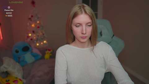 Media: Video of a young woman with fair skin and straight blonde hair, wearing a white long-sleeved top, standing in a cozy, softly lit room with a Christmas tree, plush toys, and a plush chair.