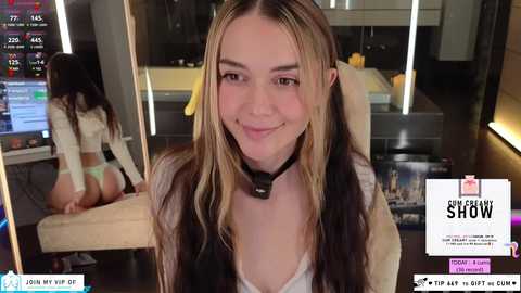 Media: Video of a smiling young woman with long, wavy blonde hair, wearing a white crop top and a black choker, sitting in a modern, dimly-lit room with a large mirror behind her.