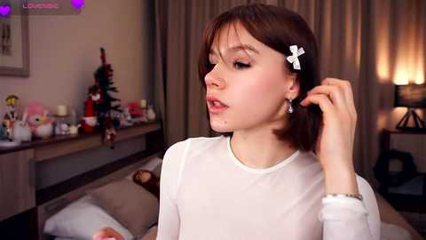 Media: Video of a young woman with fair skin, brown hair, and a white bow, holding her hair, in a cozy living room with Christmas decorations, candles, and a fireplace.