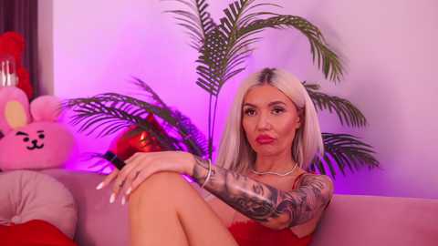 Media: Video of a blonde woman with tattoos, wearing a red dress, sitting on a couch, surrounded by plush toys and a palm tree, with a purple light background.