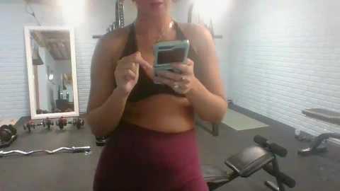 Media: Video of a woman with light skin and short, dark hair, wearing a black sports bra and maroon leggings, taking a selfie in a gym with white brick walls, equipment, and mirrors.