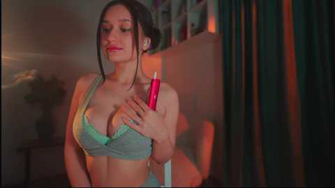 Media: A video of a young woman with fair skin, dark hair, and a curvaceous figure, wearing a green lace bra, holding a lit red candle, in a dimly lit room with green curtains.