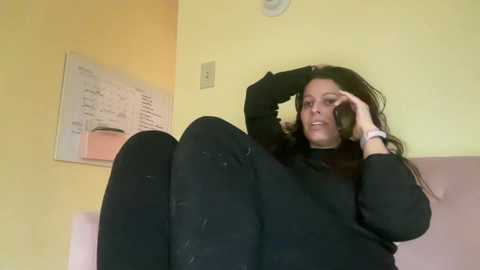 Media: A video of a woman with long black hair, wearing a black sweater and black pants, sitting on a pink couch, holding a phone to her ear, in a room with yellow walls and a whiteboard with writing.