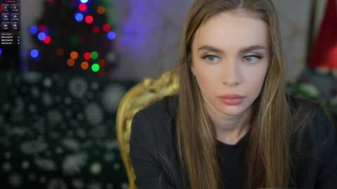 Media: A video of a young Caucasian woman with long, straight, light brown hair, blue eyes, and fair skin, wearing a black jacket, seated indoors. The blurred background features a festive Christmas tree with colorful lights.