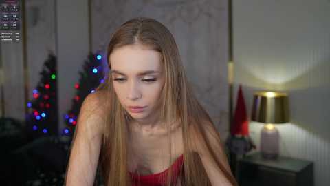 Media: A video of a young woman with long, straight blonde hair, wearing a red, sequined dress, leaning forward in a dimly lit room with Christmas decorations and a lamp.