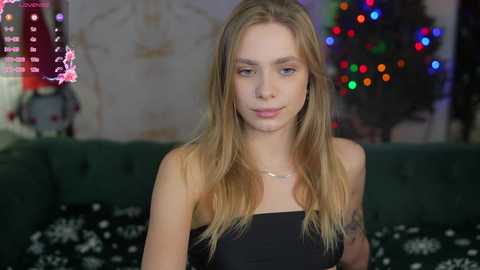 Media: Video of a young, fair-skinned woman with long blonde hair, wearing a strapless black dress, sitting on a green sofa with a festive Christmas tree and multicolored lights in the background.