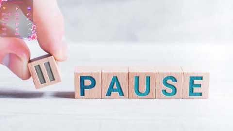 Media: Video of a hand holding a wooden block with the word \"PAUSE\" in blue letters, set on a light-colored, textured surface. The background is softly blurred, emphasizing the simplicity and minimalist design.