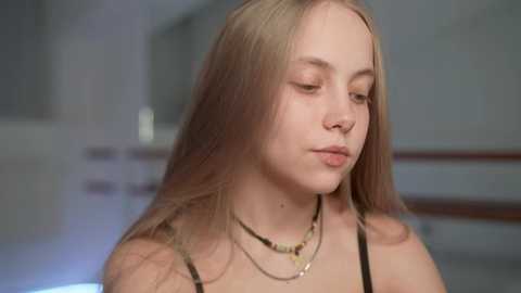 Media: Video of a young, fair-skinned girl with long, straight, blonde hair, wearing a black spaghetti-strap top and multiple layered necklaces, looking introspective in a blurred, neutral-toned background.