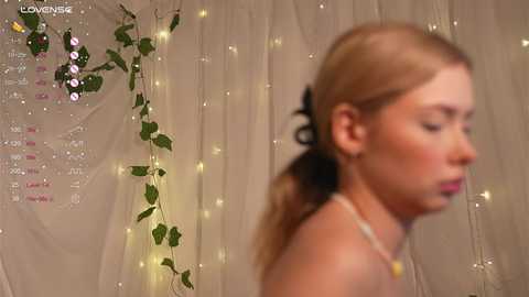 Media: Video of a blonde woman with a ponytail, wearing a necklace, standing in front of a beige curtain adorned with ivy and fairy lights.