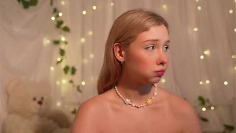 Video of a young, fair-skinned girl with straight blonde hair, wearing a white pearl necklace, looking pensive against a softly lit, white draped background with fairy lights and a teddy bear.