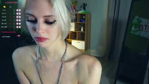 A video of a pale, slender, topless woman with platinum blonde hair, wearing a silver chain necklace, in a dimly lit room, with a TV showing a live stream.