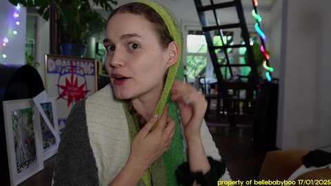 Media: Video of a young woman with short brown hair, wearing a green headscarf and a grey sweater, standing in a cozy room with a spiral staircase, colorful lights, and framed photos on the wall.