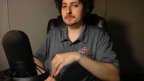 Media: Video of a man with curly hair and a mustache, wearing a blue polo shirt with a red and blue emblem, sitting in front of a microphone on a black chair against a plain background.