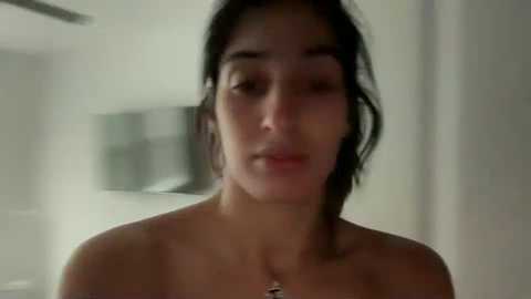 Media: A blurry video of a young woman with dark hair, fair skin, and a neutral expression. She appears topless, and the background is indistinct.