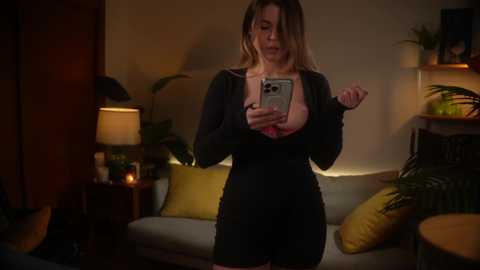 Media: Video of a blonde woman with a curvy figure, wearing a black top revealing a pink bra, taking a selfie in a dimly lit living room with green plants, yellow pillows, and wooden furniture.