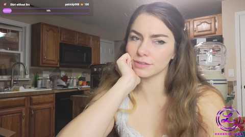 Media: Video of a young woman with long brown hair, light skin, and a slender build, resting her chin on her hand, in a modern kitchen. She wears a white tank top.