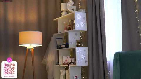 Media: Video of a cozy, modern living room with a white shelving unit filled with decorative items, a beige lamp, and fairy lights, beside a green upholstered chair.