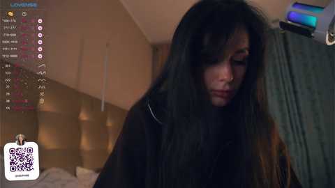 Media: Video of a woman with long black hair, wearing a black jacket, in a dimly lit bedroom, with a headboard and a green curtain visible.