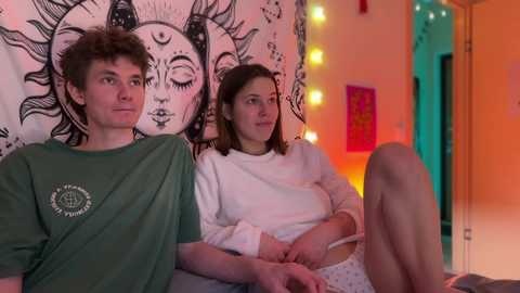 Video of a young couple sitting on a bed in a cozy, colorful room with tapestries, fairy lights, and a relaxed atmosphere.