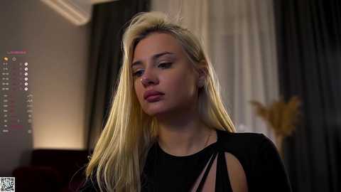 A video of a blonde woman with straight hair, wearing a black dress, looking thoughtful, in a dimly lit room with curtains and a TV screen.