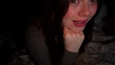 Video of a young woman with long dark hair, light skin, and flushed cheeks, resting her chin on her hand, looking pensive. She wears a green top. Background is dimly lit with a dark, textured surface.