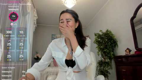 Media: Video of a young Asian woman with long dark hair, wearing a white shirt tied at the waist, covering her black bra, wiping her eyes in a bedroom with a bed, dresser, and plant.