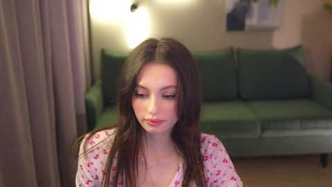 Media: Video of a young Caucasian woman with long brown hair, wearing a floral-patterned shirt, seated in a dimly-lit living room with a green couch and beige curtains in the background.