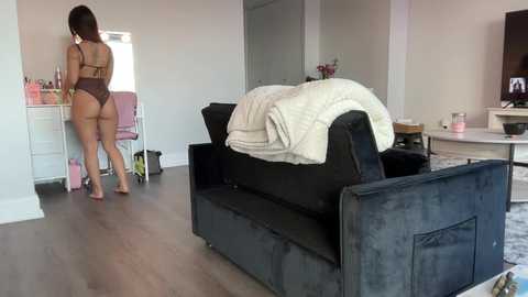 Media: Video of a young woman with short brown hair, wearing a tan thong bodysuit, standing in a modern living room with a black sofa, white bedding, and a pink chair.