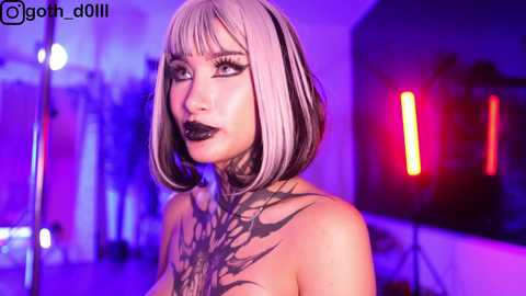 Media: Video of a woman with platinum blonde bob haircut, dark lipstick, and intricate black tattoo across chest, standing in a dimly lit room with purple and red neon lights, looking contemplative.