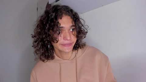 Media: Video of a young woman with curly, shoulder-length brown hair, light brown skin, and a soft smile. She wears a beige top, standing against a plain white wall.