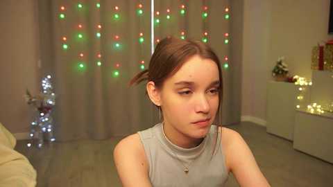 Media: Video of a young woman with light skin, brown hair tied back, wearing a gray sleeveless top, sitting in a warmly lit room with festive fairy lights and Christmas decorations in the background.