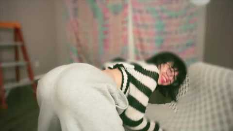 Media: A blurry video of a woman with dark hair and fair skin, wearing a black and white striped sweater and white pants, lying on a beige couch, laughing. The background features a colorful, abstract wall hanging and a wooden step ladder.