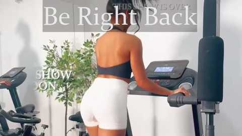 Media: Video of a fit, dark-skinned woman with long black hair, wearing a black sports bra and white shorts, using an elliptical machine in a modern gym. Background includes green plants and motivational text.