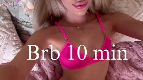 Video of a blonde woman in a bright pink bra, lying on a bed with floral pillows and a textured blanket. Text overlay reads \"Brob 10 min.\