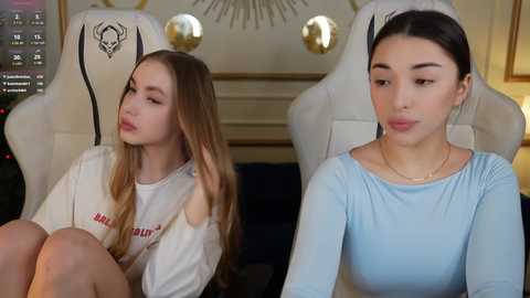 Video of two young women, one in a white \"Nasty Gal\" T-shirt, the other in a light blue long-sleeve top, seated on white gaming chairs in a modern, dimly-lit room with circular gold decorations on the wall.