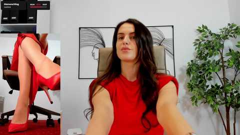 Media: Video of a woman with long brown hair, wearing a red sleeveless dress, sitting in an office chair with a plant and abstract artwork in the background.