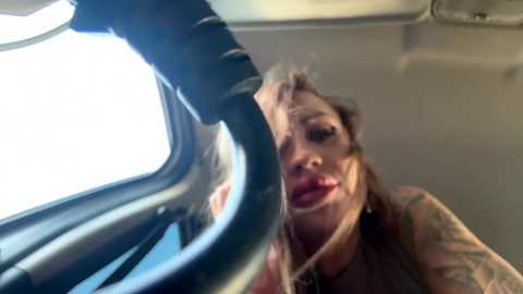 Media: Video of a woman with a tattooed arm, wearing a dark top, and large red lipstick, leaning out of a car window, with the cityscape visible outside.