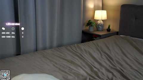 A video of a modern bedroom with a gray upholstered headboard, a beige bedspread, a dark wooden nightstand with a lamp, and green plants, featuring a \"Live View\" QR code in the bottom left corner.