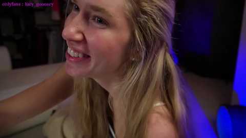 Media: A video of a smiling, blonde, fair-skinned young woman with braces, wearing a light-colored top, in a dimly lit room with a blue glow.