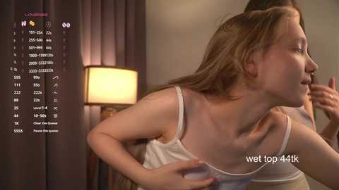 Media: A video depicts a young woman with light skin and straight, shoulder-length blonde hair, wearing a white tank top, leaning forward with her hands on her chest. A cozy room with a warm, yellow lamp and soft curtains is in the background.