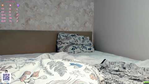 Media: Video of a messy, unmade bed with floral-patterned white sheets, two pillows, and scattered clothes. The room has a beige headboard and a textured, light-colored wall. A smartphone lies on the bed.