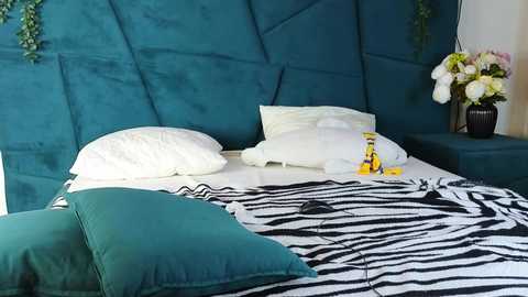 Media: Video of a modern bedroom featuring a teal tufted headboard, white pillows, zebra print duvet, green and teal pillows, yellow toy car, and a vase of flowers on a nightstand.