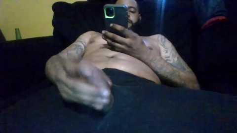 Media: Video of a muscular, tattooed man with light skin, shirtless, holding a smartphone, lying on a dark couch, wearing black pants, indoors with dim lighting.