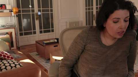 Media: Video of a curvy, dark-haired woman with medium skin tone in a cozy living room, wearing a brown sweater, leaning on a chair, with glass doors and wooden furniture in the background.